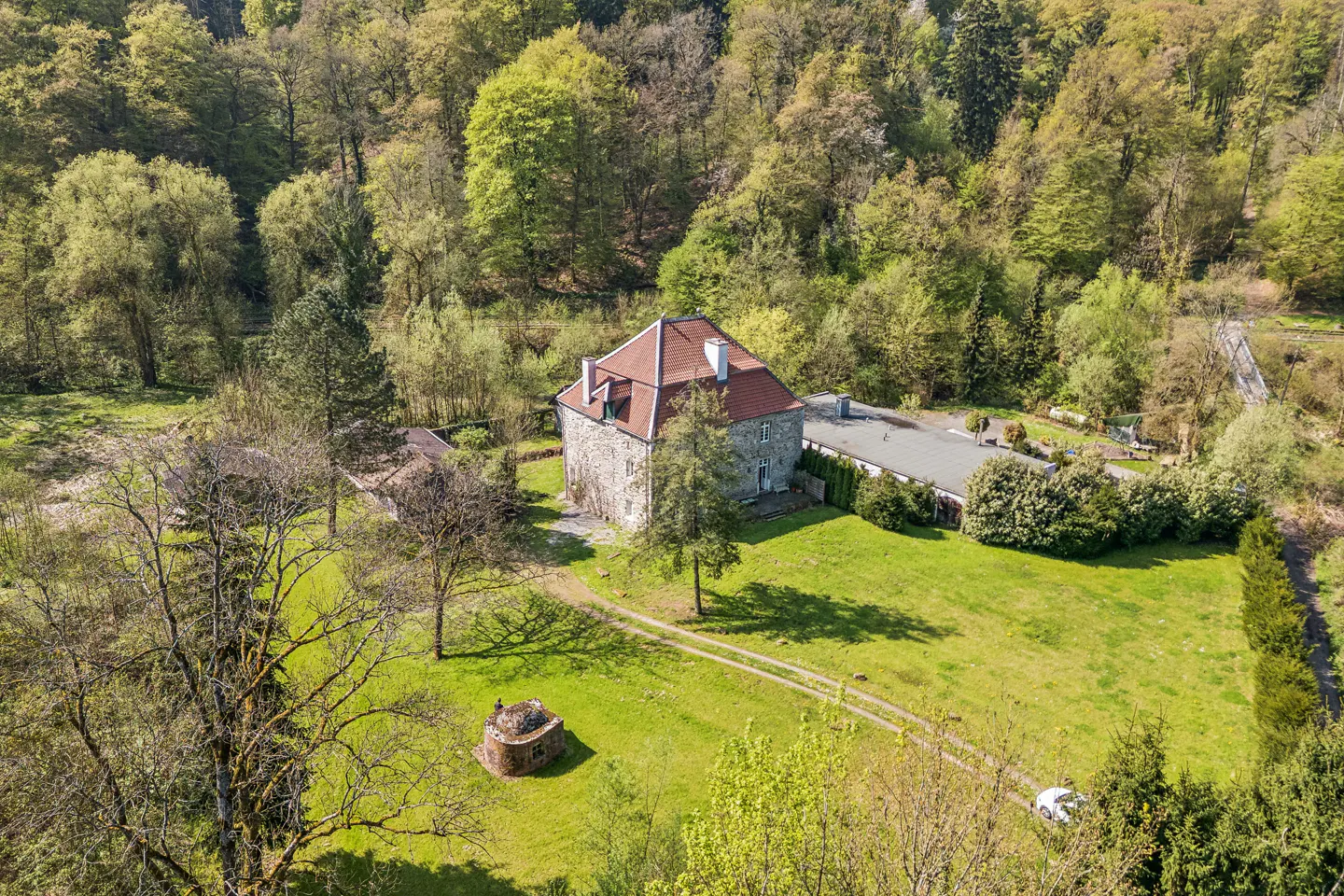 Aerial view of a stone house with a red tile roof, surrounded by green lawn and trees. A small stone structure sits near a dirt path.