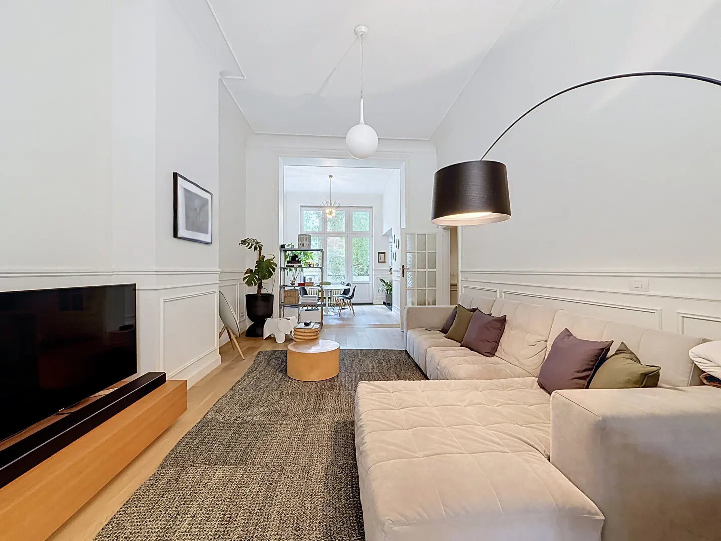 Modern living room with a large beige sectional, dark area rug, TV, and an arched floor lamp, leading to a dining area.