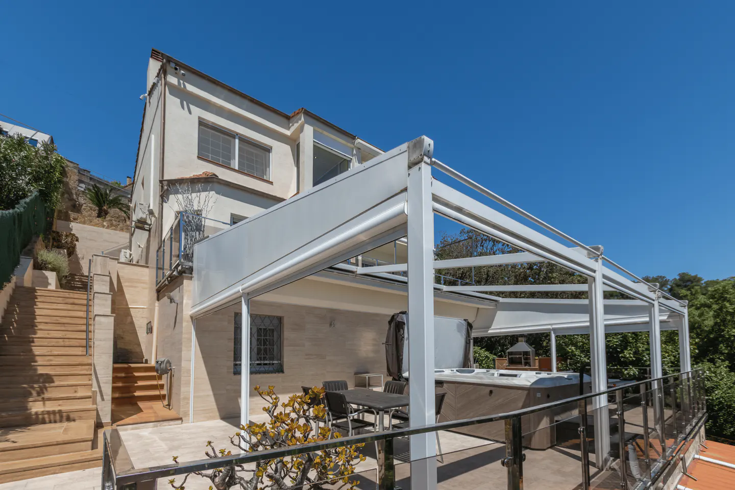Exterior view of a two-story house with a patio, hot tub, table, and chairs under a white pergola. Stairs lead up the hillside.