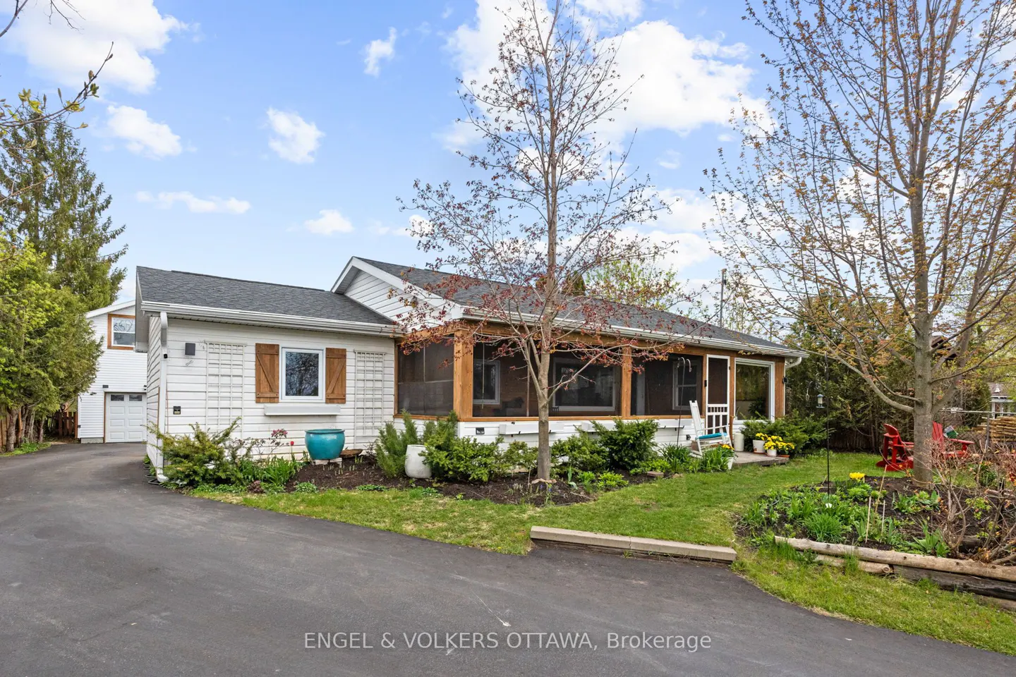 Exterior view of a one-story white house with a screened-in porch, black roof, and a driveway. The house is surrounded by greenery and trees.