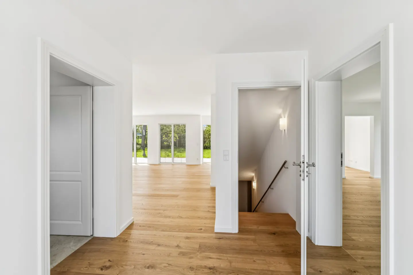 Bright hallway with light wood floors, white walls, multiple doorways, and a staircase leading to a lower level.