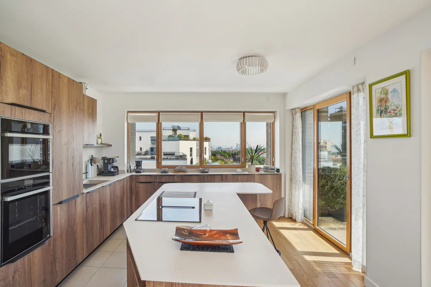 Bright kitchen with wood cabinets, white countertops, and island. Large windows offer city views. A sliding glass door leads to a balcony.