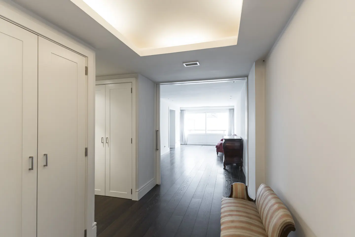 Hallway with white walls, dark wood floors, and white closets. A striped bench sits on the right, leading to a bright living room.