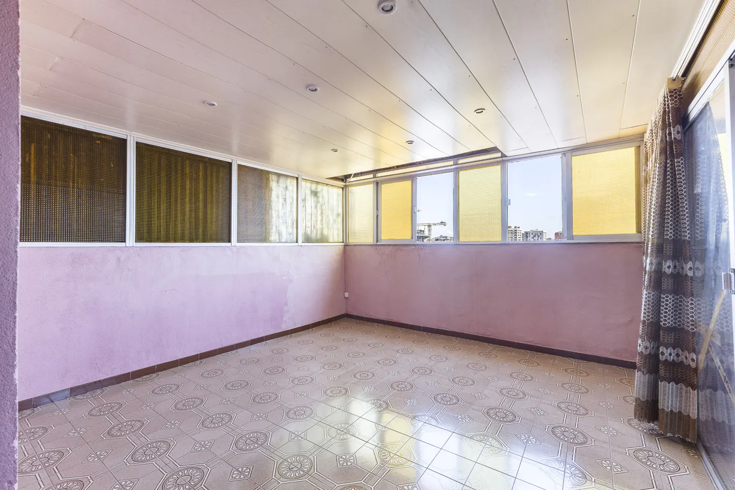 Empty sunroom with pink walls, patterned tile flooring, and large windows with yellow-tinted glass and patterned curtains.