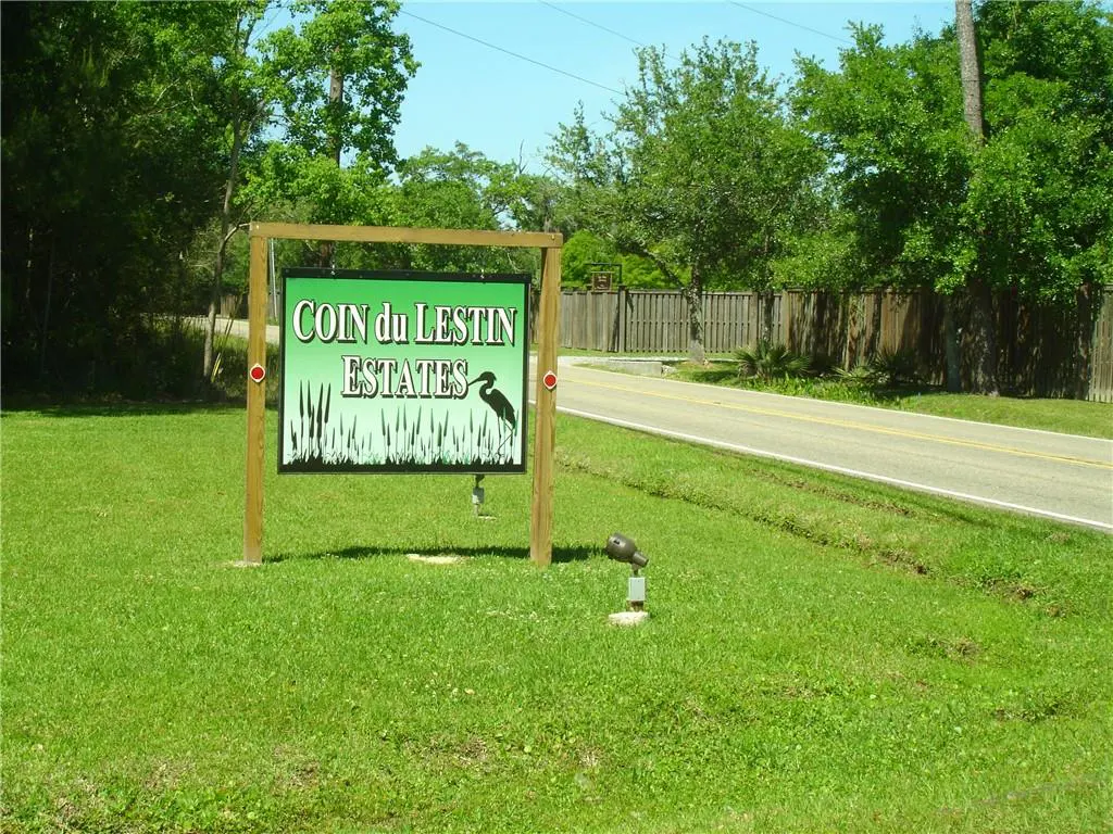 "Coin du Lestin Estates" sign on green lawn. Sign has heron and reeds graphic. Road and trees in background.