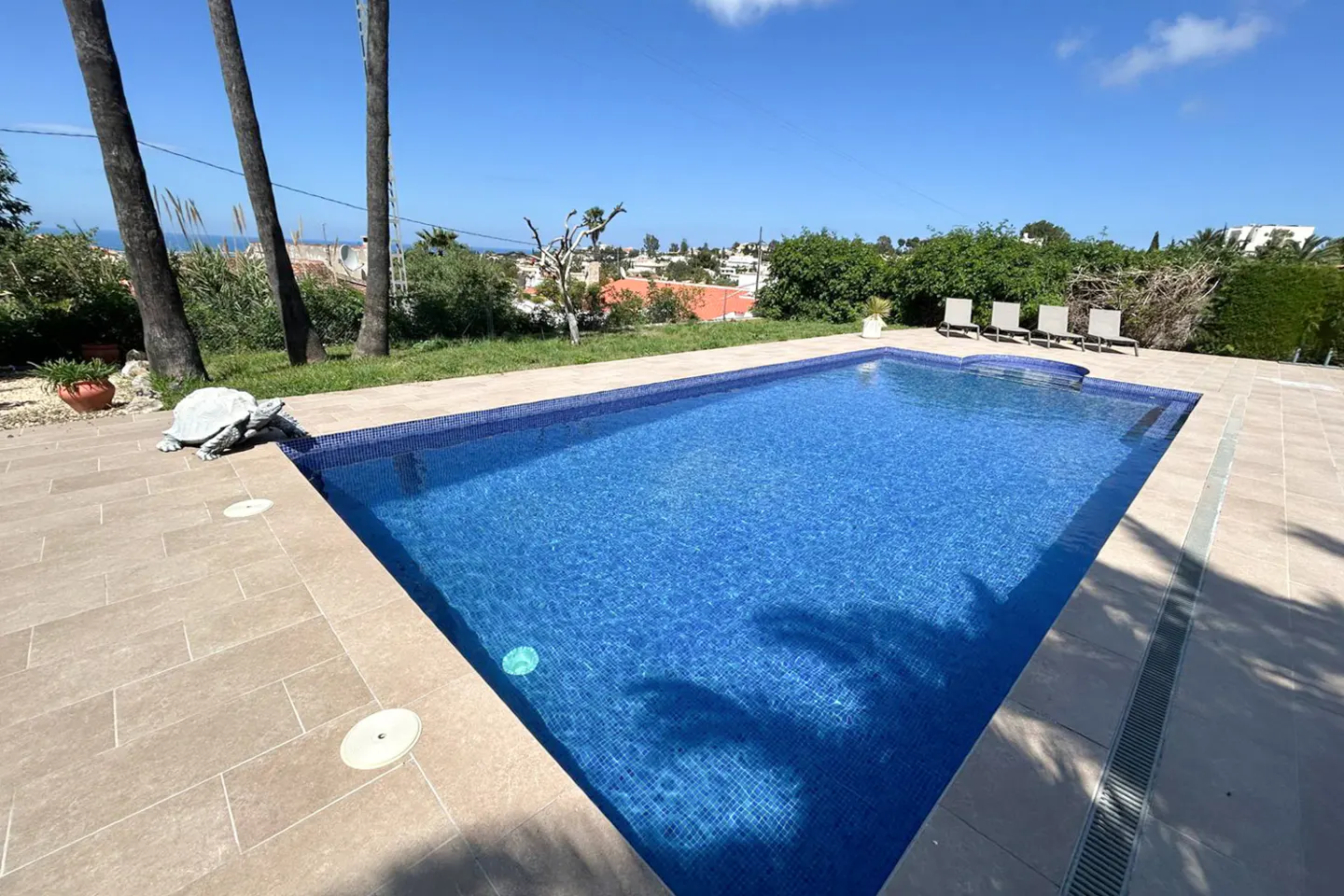 A blue tiled swimming pool is surrounded by tan pavers, with palm trees and a turtle statue nearby.