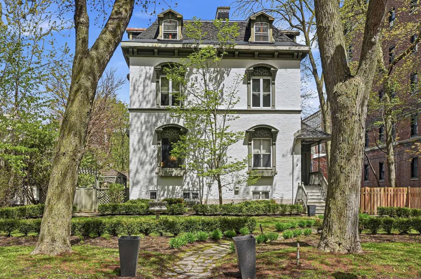 A three-story white brick house with a black roof and dormer windows, framed by trees and a stone path.