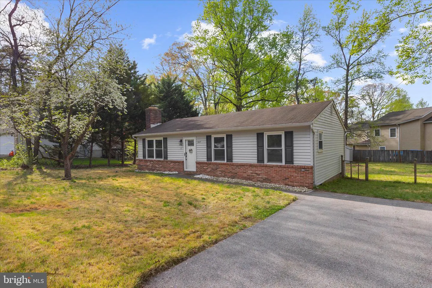 A single-story house with white siding, a brick foundation, and black shutters. A gray driveway leads to the house, surrounded by trees and a lawn.