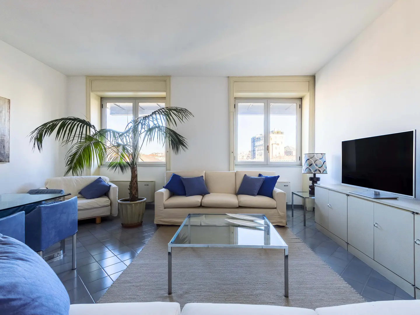 Bright living room with white walls, gray tile floor, and large windows. Beige sofa with blue pillows, glass coffee table, and a large potted palm.
