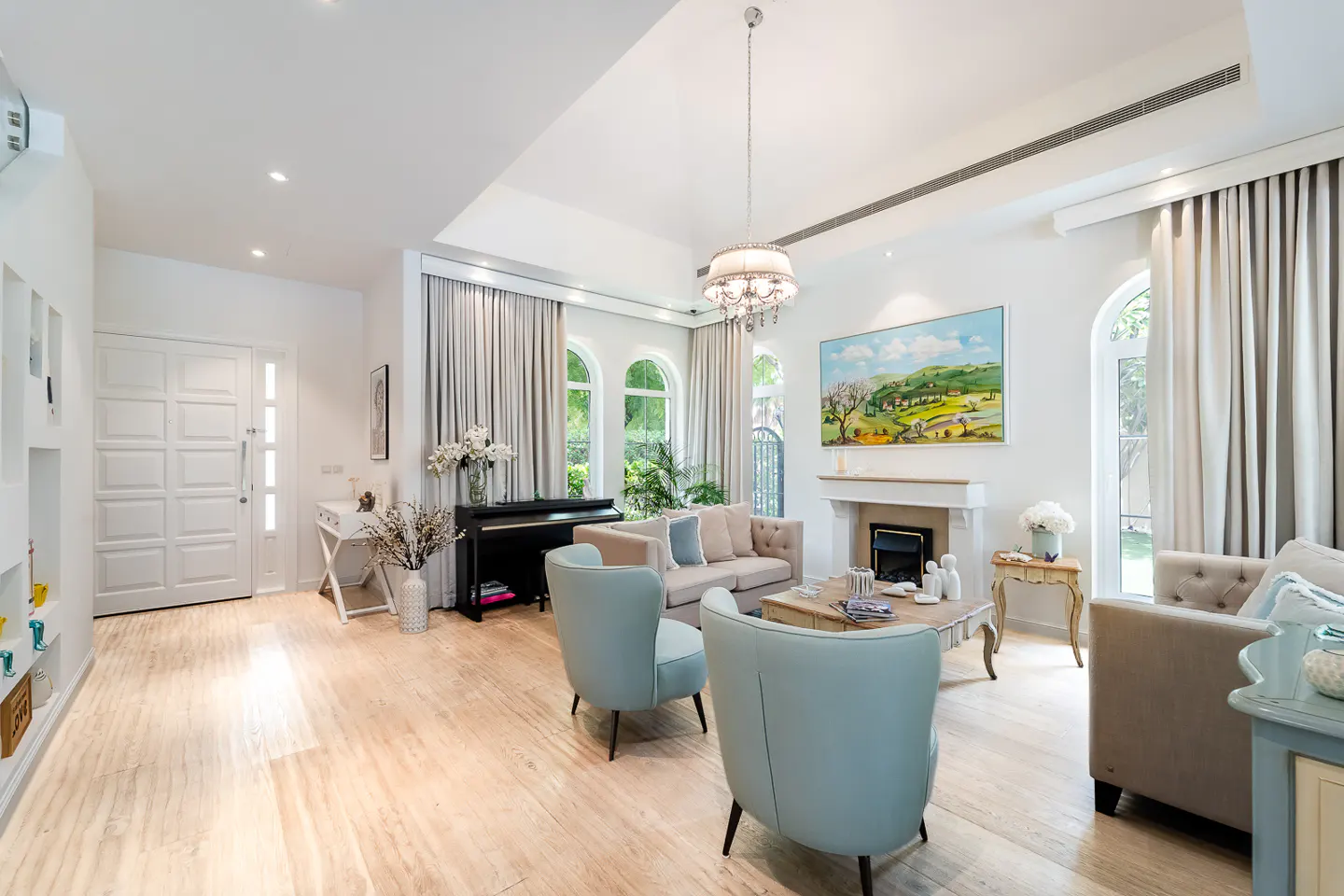 Bright living room with light wood floors, white walls, and high ceilings. Features a piano, fireplace, sofas, and blue armchairs.