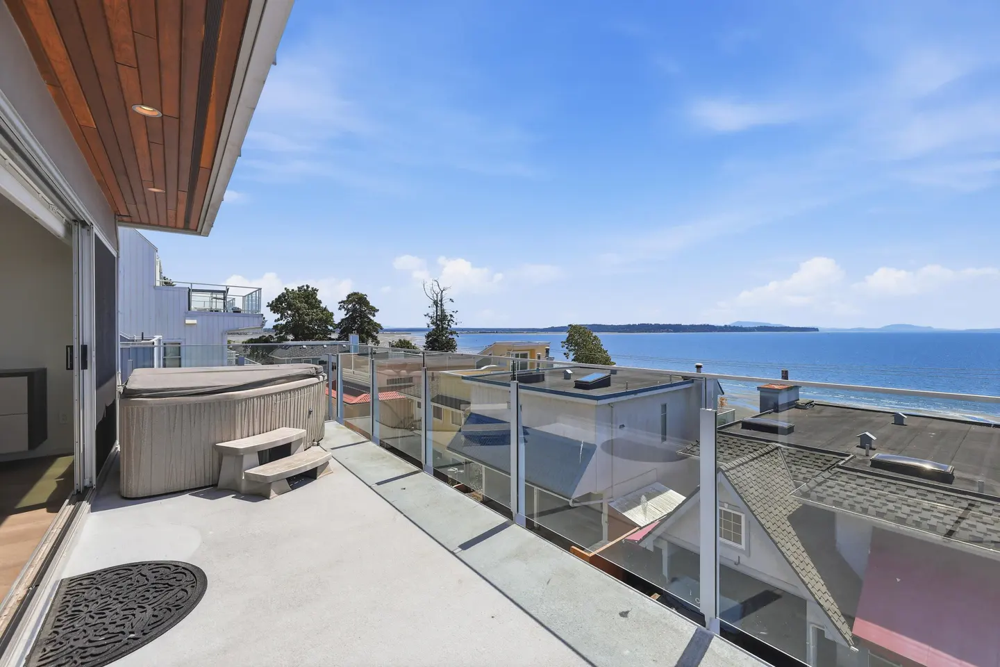 Modern balcony with a hot tub, glass railing, and panoramic ocean views under a clear blue sky.
