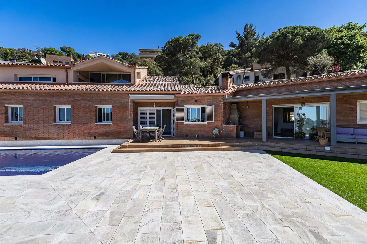 Brick house with a gray tiled patio, pool, and green lawn. Trees and other houses are in the background.