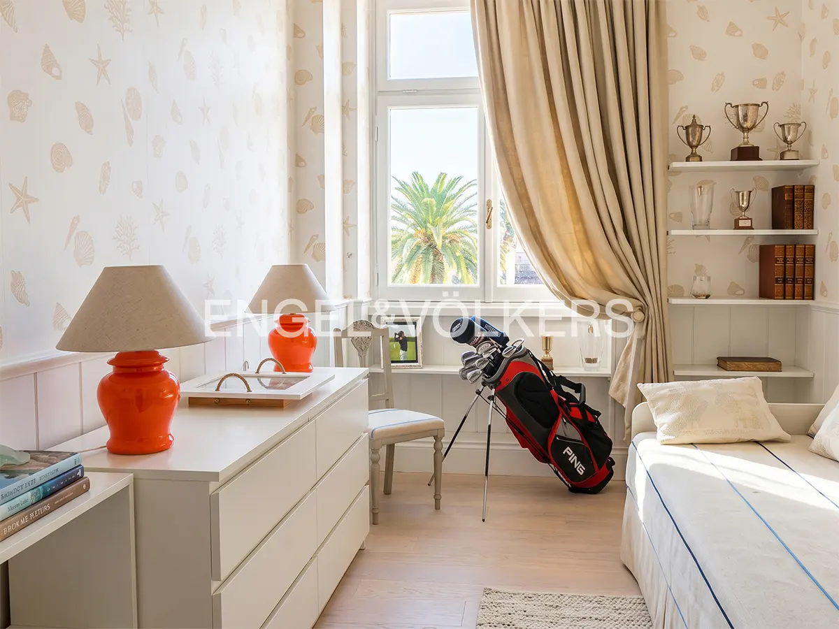 Bedroom with seashell wallpaper, white dresser with orange lamps, a golf bag, and shelves displaying trophies and books.