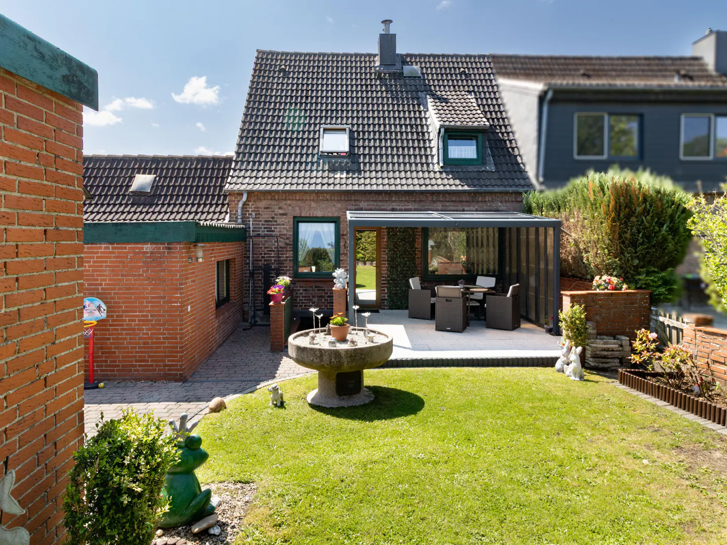 A brick house with a green lawn, stone fountain, and covered patio with outdoor furniture.