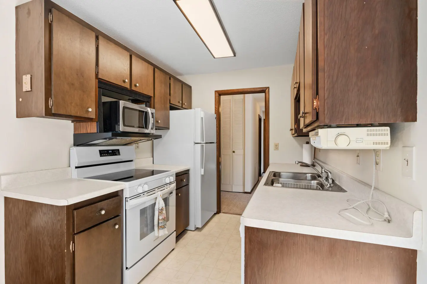 A kitchen with brown cabinets, white appliances, and light countertops. A microwave is above the stove. A doorway is visible in the background.