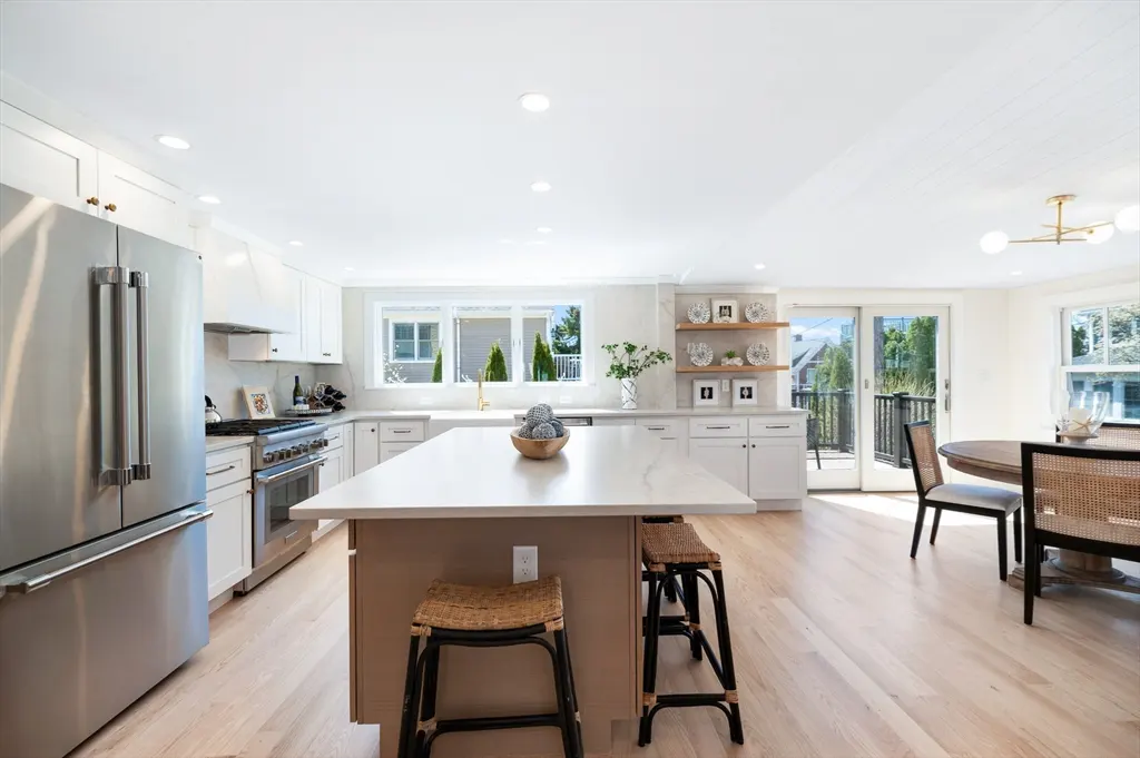 Bright, modern kitchen with white cabinets, stainless steel appliances, and a large island with seating. A dining area is visible in the background.