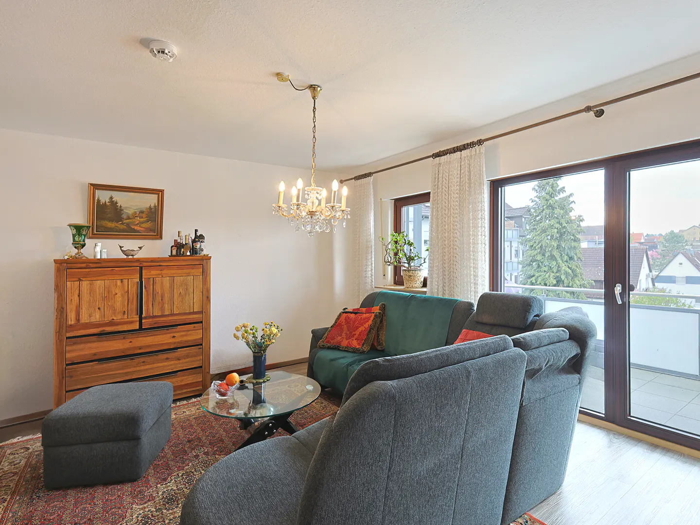 Living room with gray sofas, green loveseat, and wooden cabinet. A chandelier hangs above a glass coffee table on a patterned rug. Balcony doors show a view.