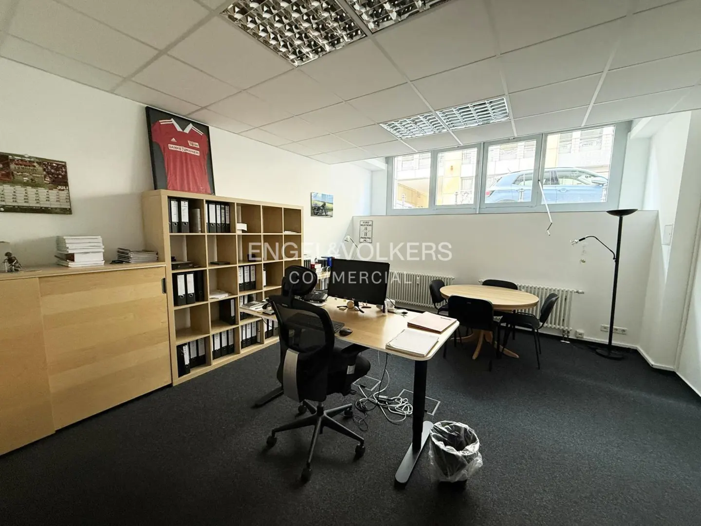 Office space with a desk, chair, and computer. A bookshelf with binders and a framed jersey are visible. A round table with chairs is in the background.