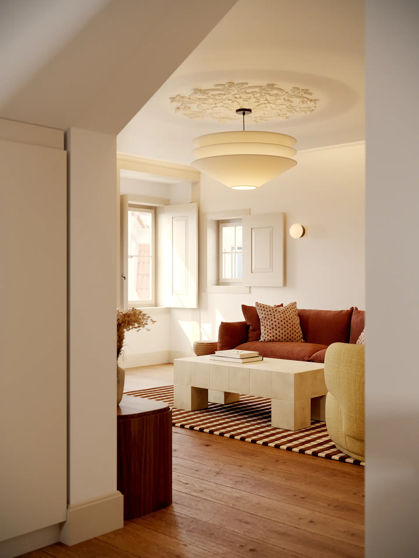 Bright living room with wood floors, a brown sofa, and a large white coffee table. A striped rug sits beneath the furniture. Windows with white shutters let in natural light.
