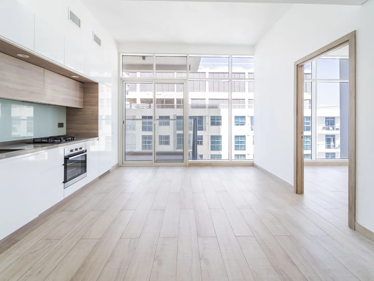 Bright, empty apartment with white walls, light wood floors, and a modern kitchen. Large windows offer a city view.