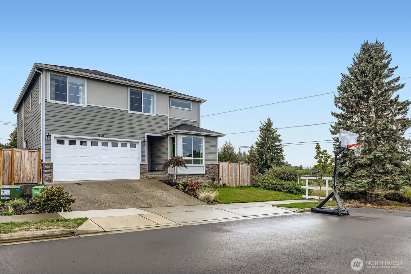 Two-story gray house with a white garage, concrete driveway, and a basketball hoop in the front yard under a blue sky.