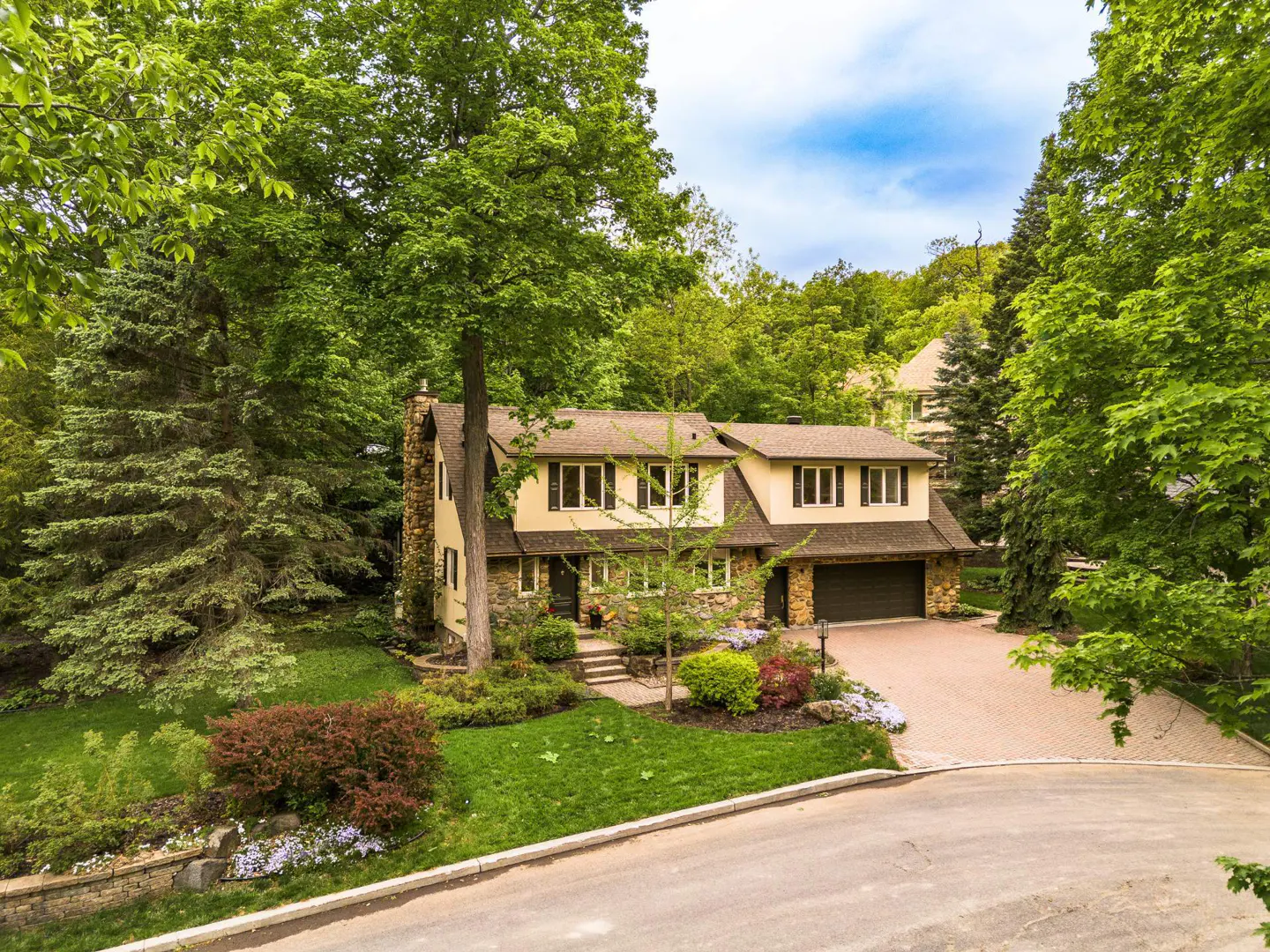 Two-story home with stone and tan siding, a paved driveway, and lush green landscaping in a wooded setting.