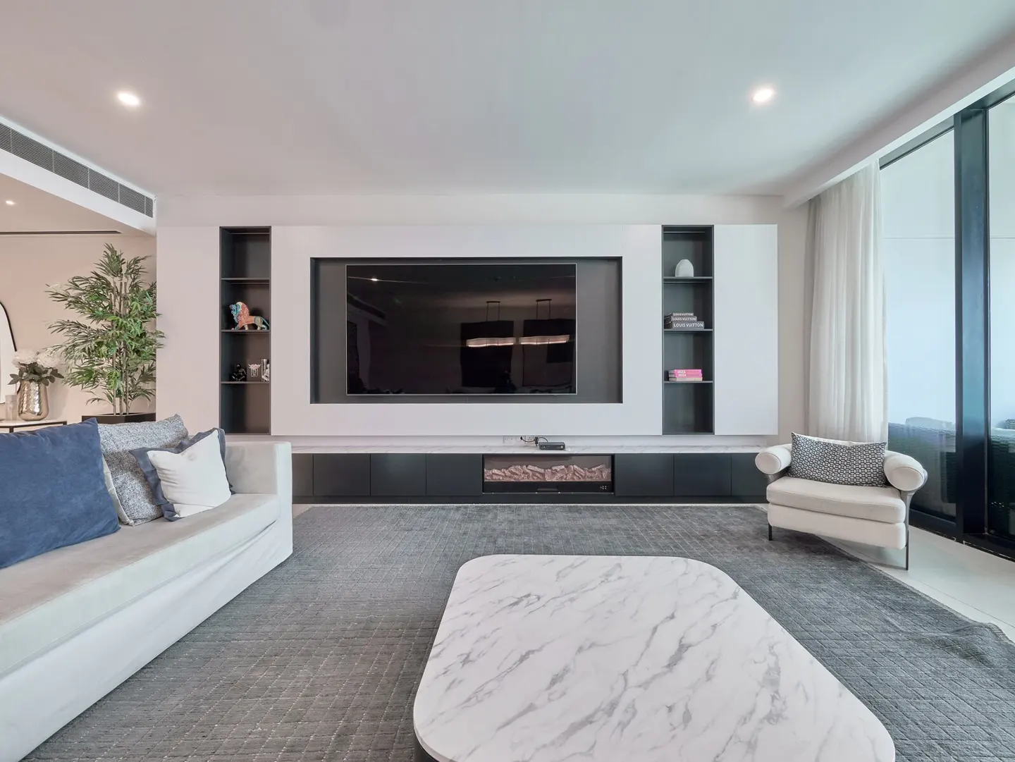 Living room with a white sofa, marble coffee table, and a large TV mounted on a white wall unit with black shelves.