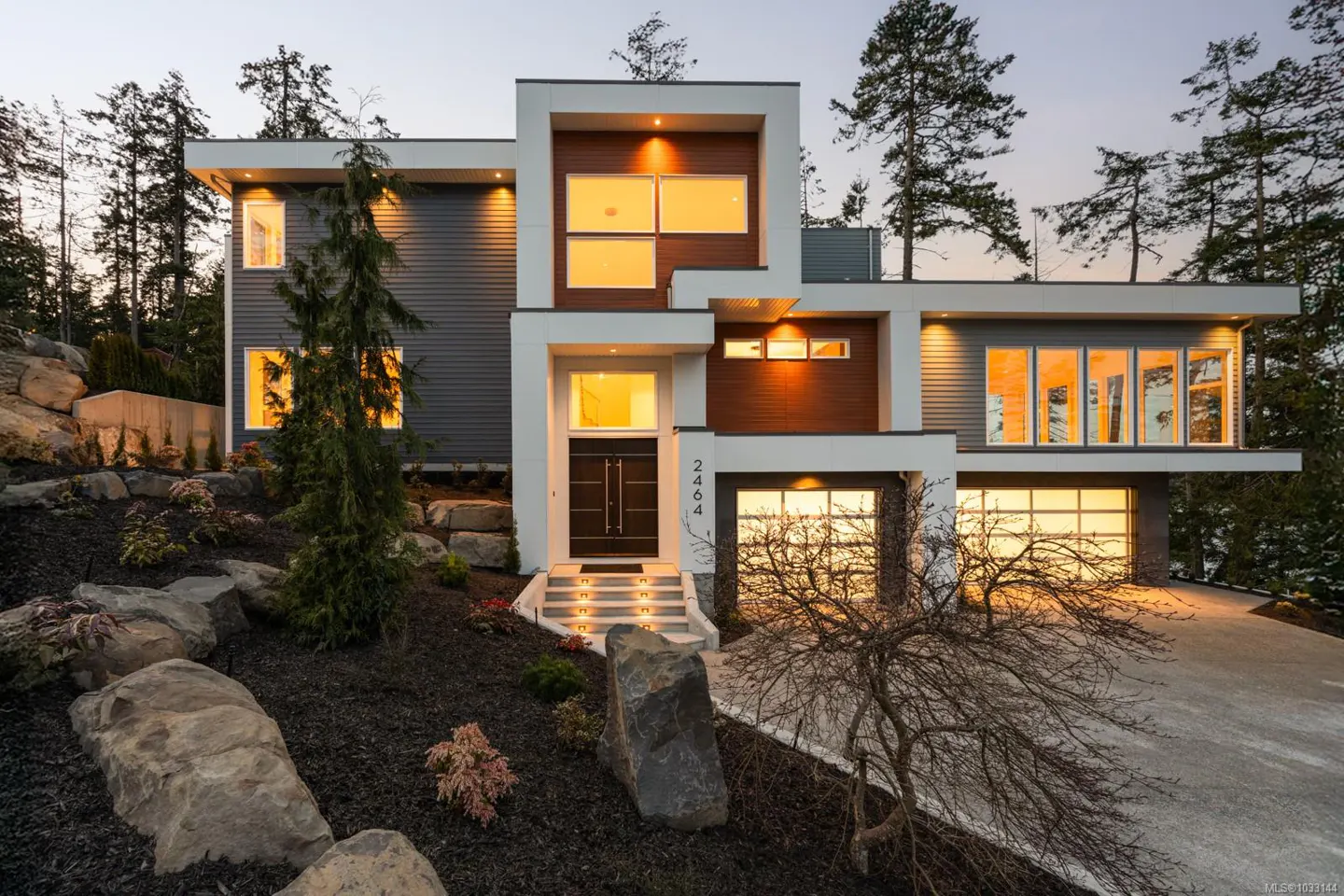 Modern two-story house with gray siding, white trim, and wood accents. Large windows glow with warm light. Landscaped yard with rocks and plants.