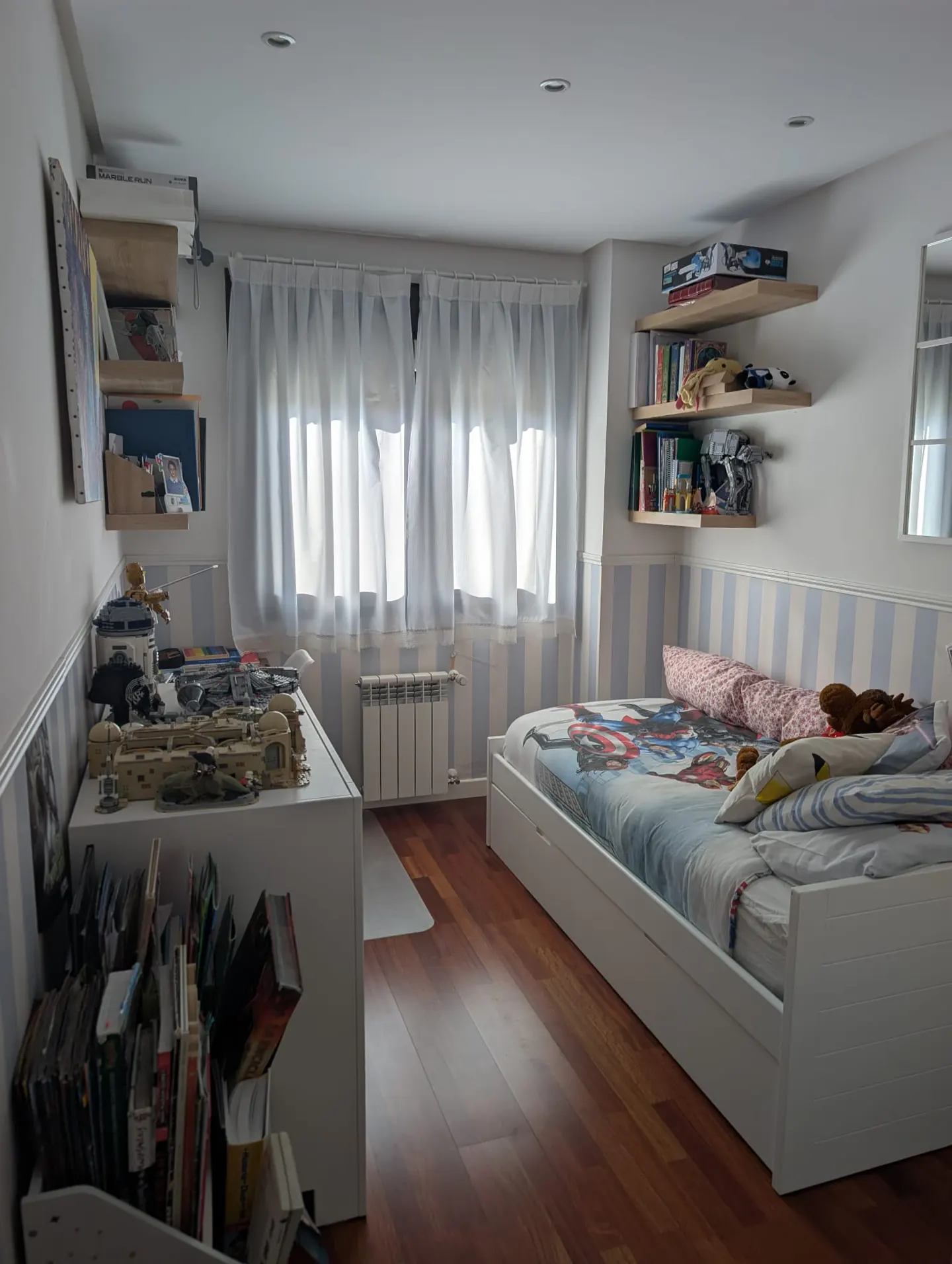 A child's bedroom with a bed, white desk, wooden shelves, and light blue striped wallpaper, featuring a window with curtains.