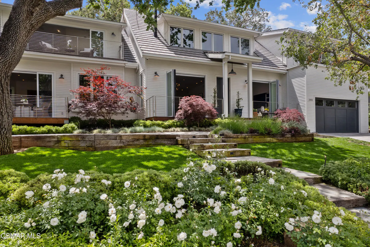 Two-story white house with a gray roof, dark gray garage door, and a green lawn with white flowers.