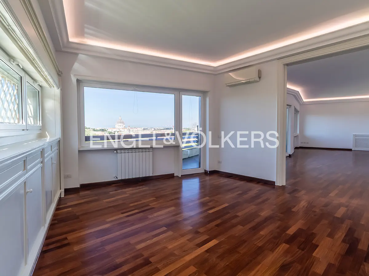 Bright, empty room with hardwood floors, white walls, and large windows with a city view. Another room is visible through a doorway.