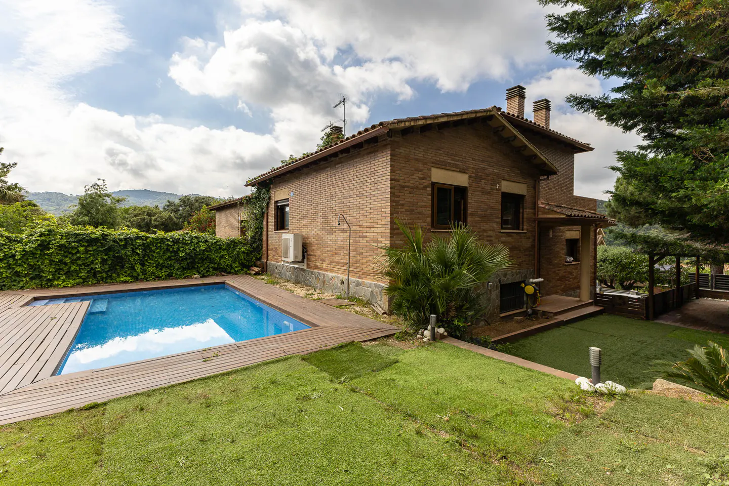 Brick house with a blue pool and wooden deck, surrounded by green grass and trees under a cloudy sky.