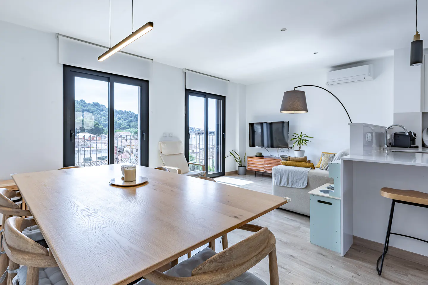 Modern open-plan living area with a light wood dining table, white sofa, TV, and large glass doors overlooking a hillside.