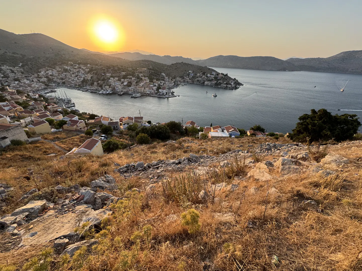 Sunset over a Greek coastal town with white houses, a harbor with boats, and rocky hills overlooking the calm blue sea.