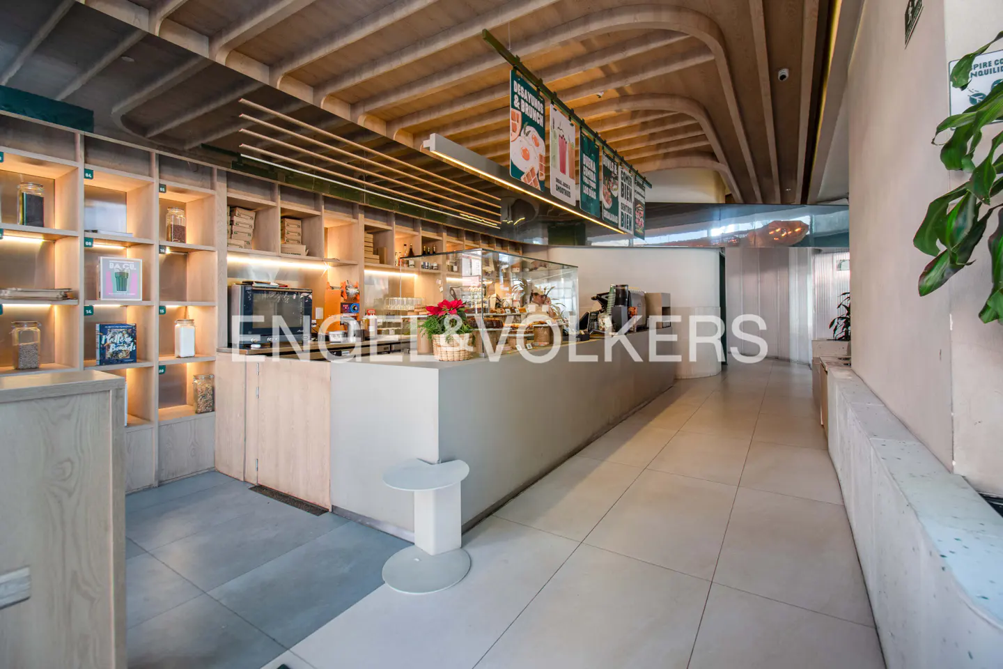 Interior view of a cafe with a counter, shelves, and a concrete ceiling. The floor is tiled and there are plants in the background.