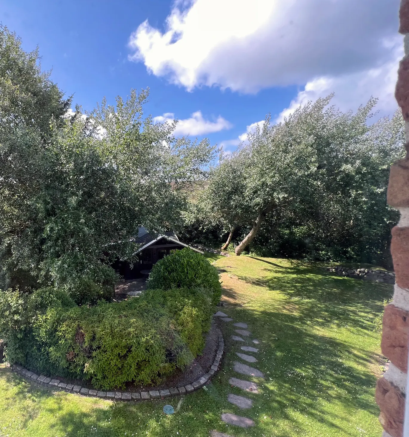 View of a lush green backyard with trees, a stone path, and a small shed under a blue sky with white clouds.