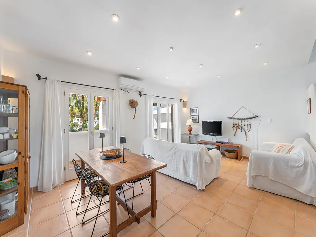 Bright living room with terracotta tile floor, white walls, and recessed lighting. A wooden table and chairs sit near a glass-front cabinet. Two white sofas face a TV.