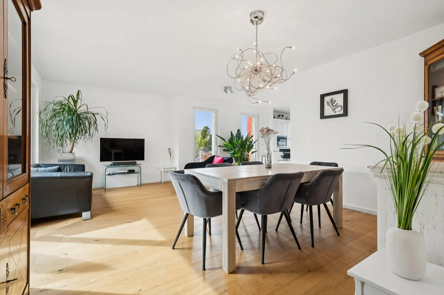 Bright, open-concept living and dining area with wood floors, white walls, and modern chandelier. Table with gray chairs, black sofa, and TV.