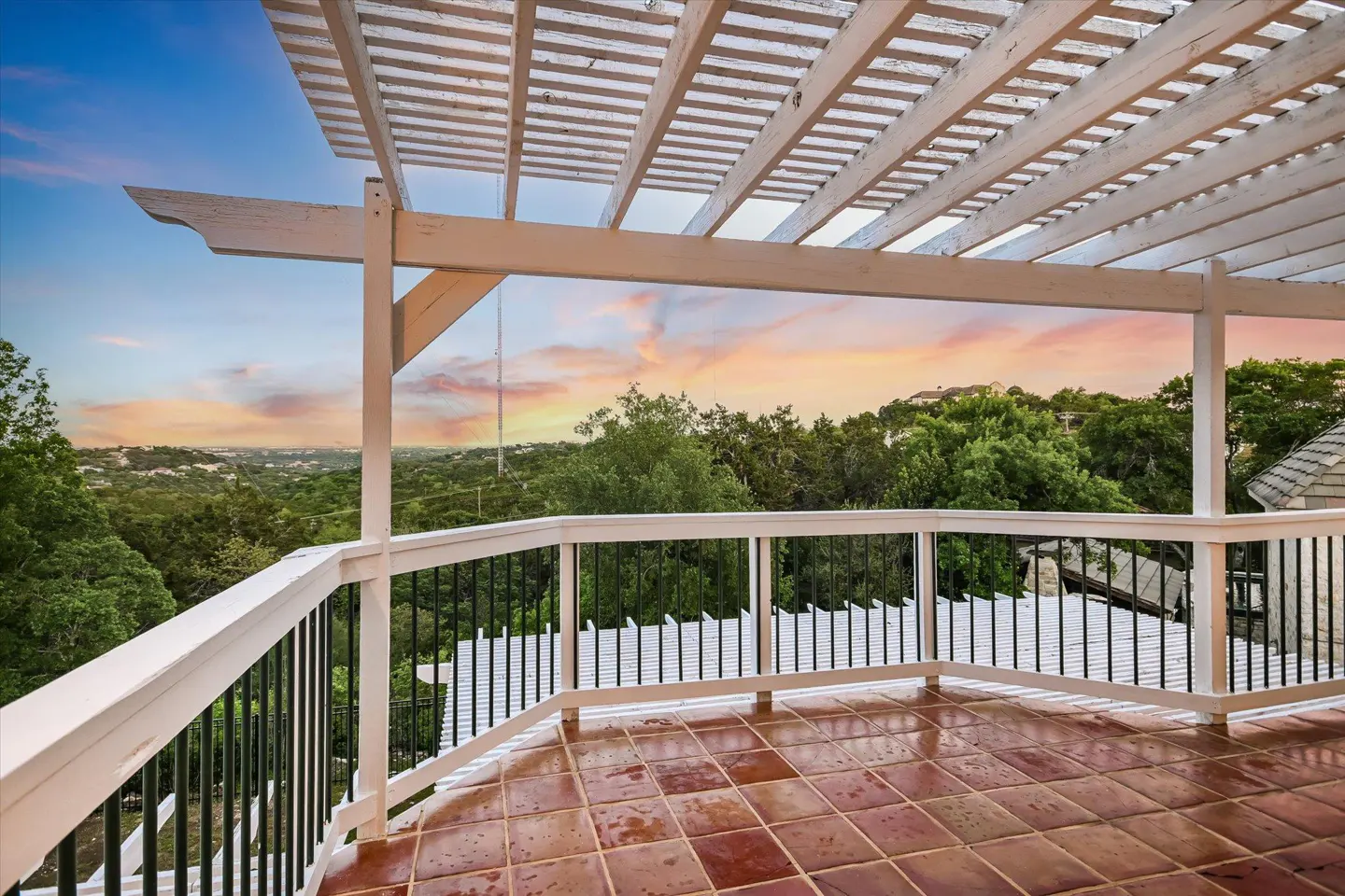 Outdoor patio with terracotta tile floor, white pergola, and black railing overlooking a green, tree-filled landscape at sunset.