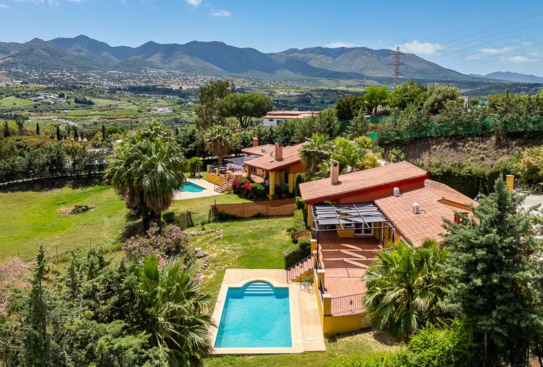 Aerial view of yellow houses with red tile roofs, pools, green lawns, trees, and mountains in the background.
