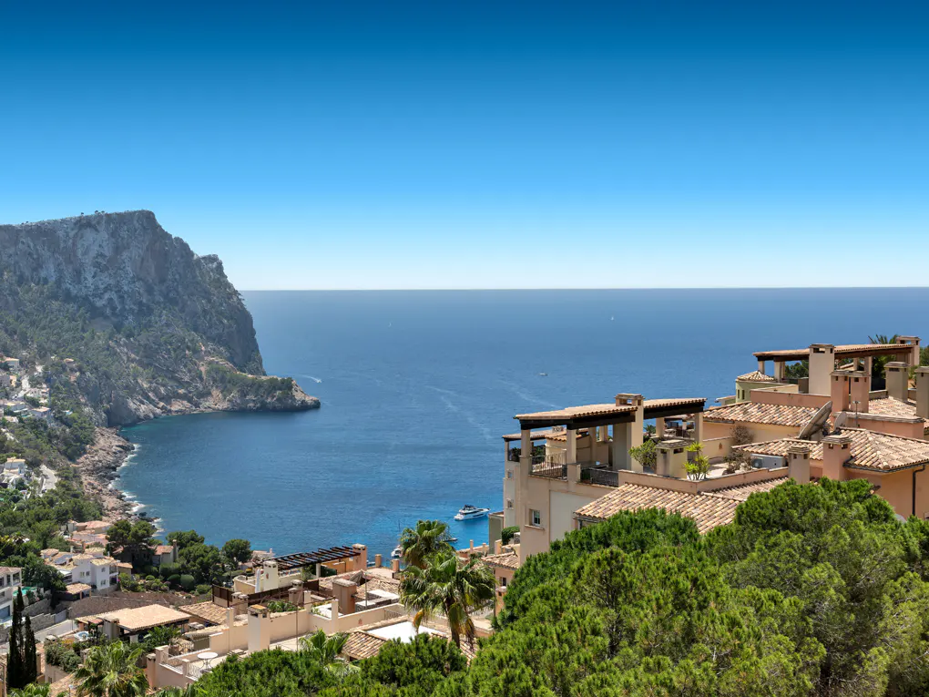 Coastal view of Mediterranean villas with terracotta roofs overlooking a blue bay, rocky cliffs, and a clear sky.