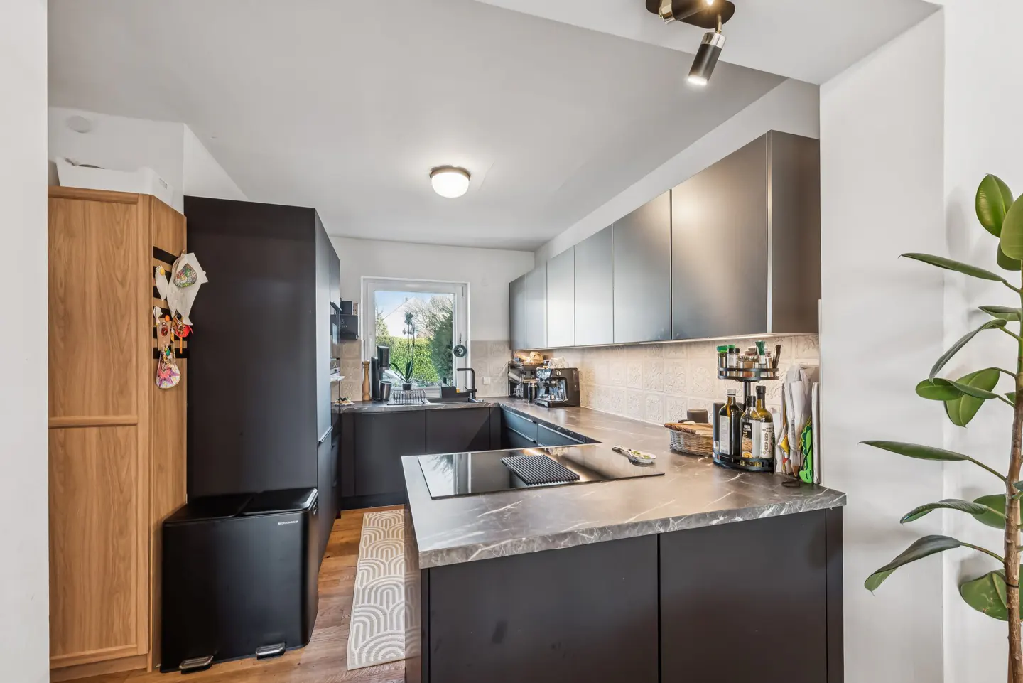 A modern kitchen with dark gray cabinets, marble countertops, and stainless steel appliances. A window overlooks a green garden.