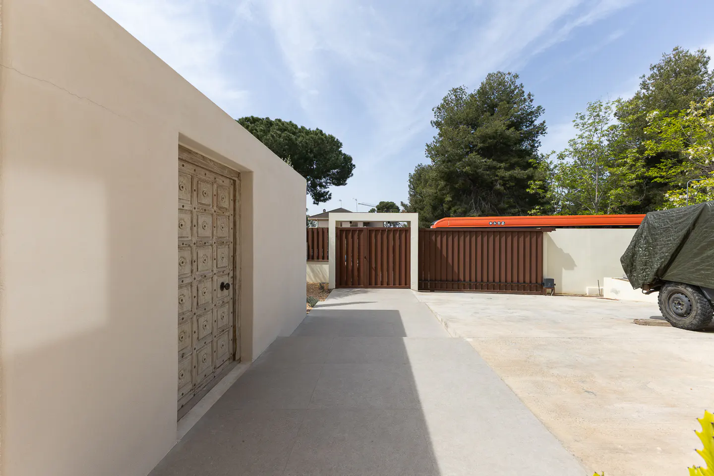 Exterior view of a beige building with a decorative wooden door, a concrete walkway, and a brown gate in the background.