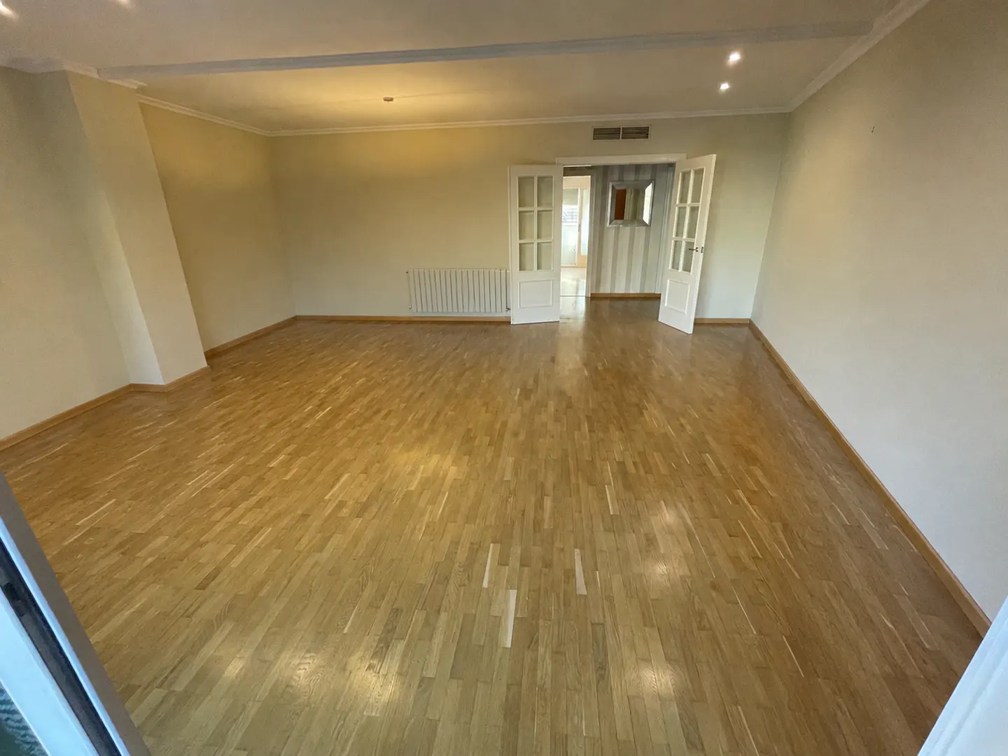 Spacious, empty room with light wood flooring, beige walls, and white double doors leading to an adjacent hallway.