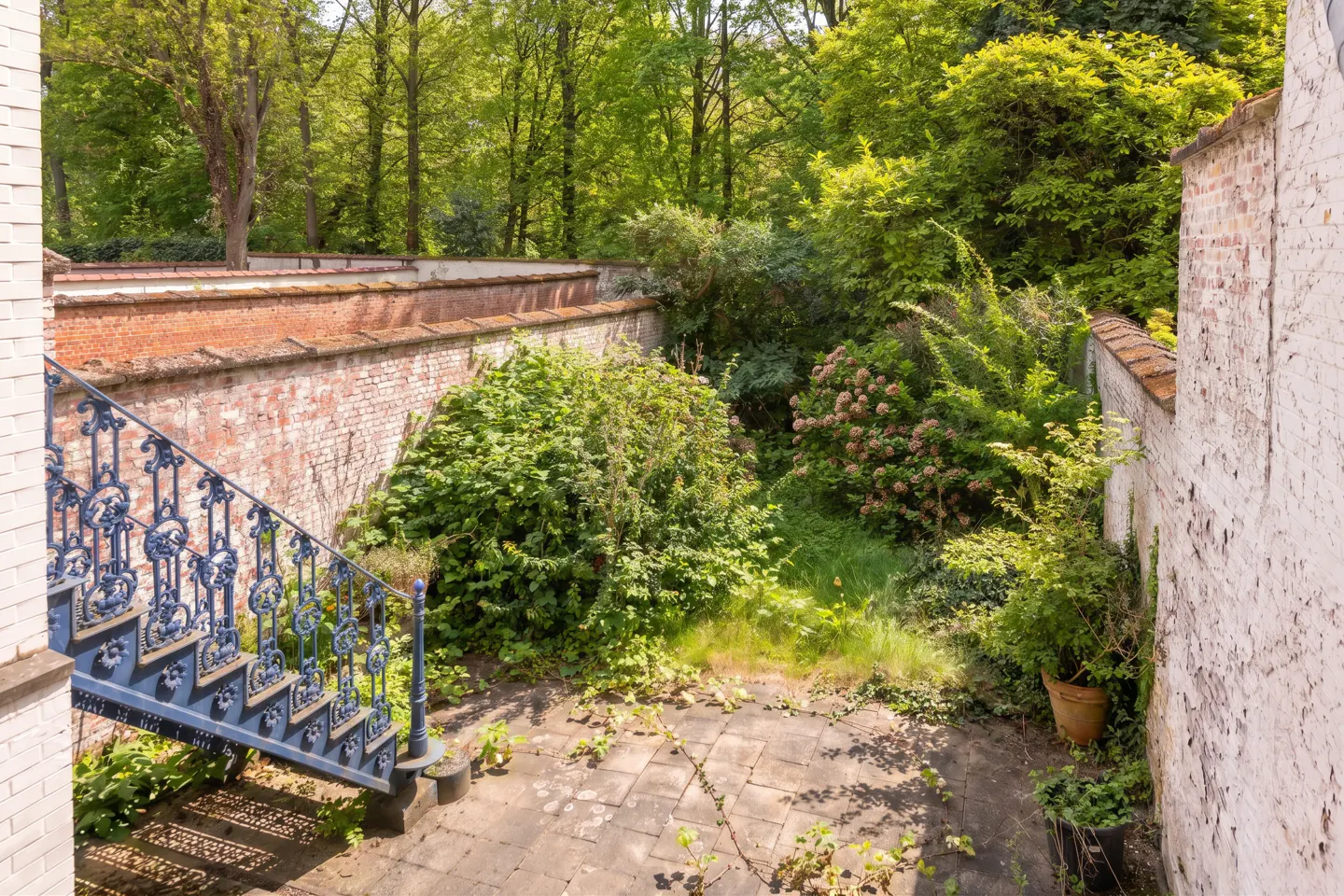 A stone patio with blue iron stairs, surrounded by brick walls and lush green trees.