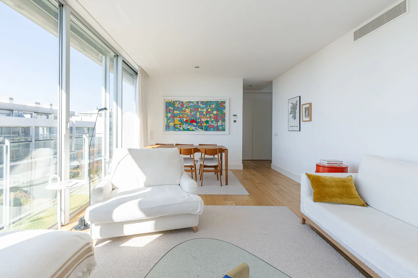 Bright, modern living room with white sofa, chair, and walls. Large windows offer city views. A wooden dining table sits near a colorful art piece.