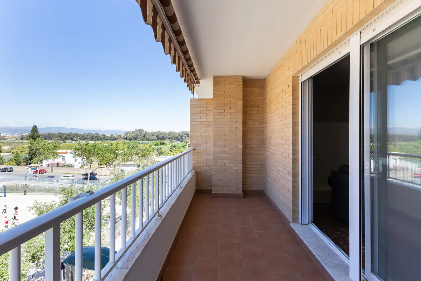 Balcony with white railing, brick wall, and brown tile floor. View of trees and blue sky. Sliding glass door to interior.