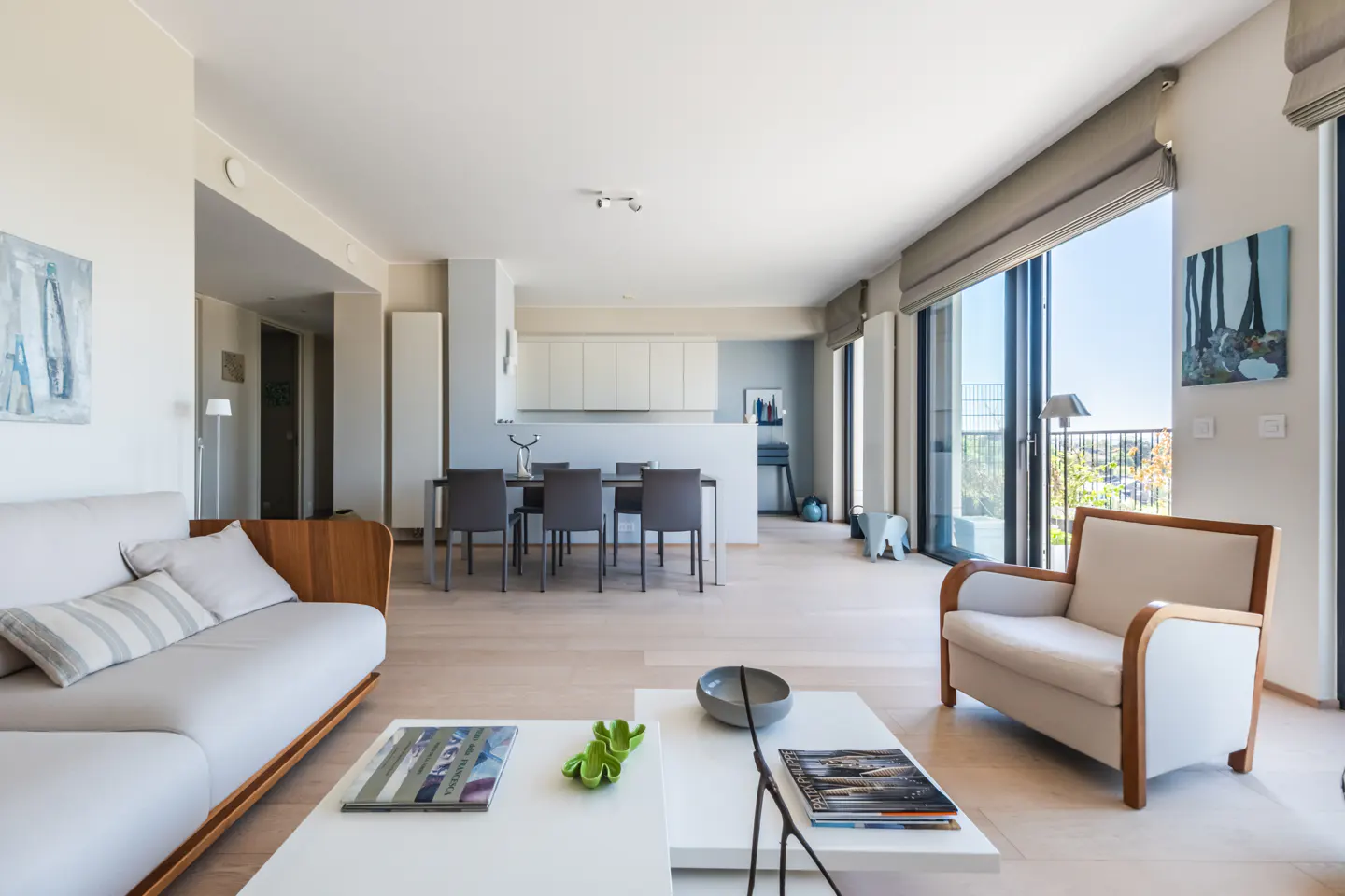 Bright, modern living room with a white sofa, armchair, and coffee tables. Dining table and kitchen in the background. Large windows with a view.