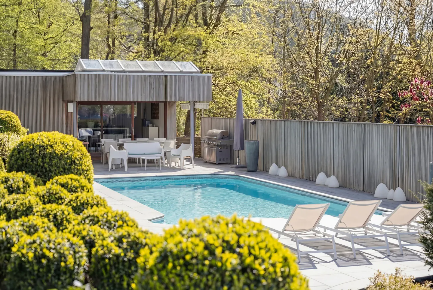 A backyard with a blue swimming pool, lounge chairs, a patio with white furniture, and green bushes.