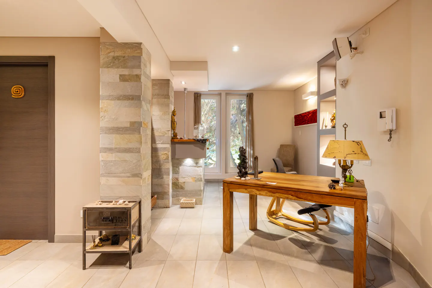 Modern home office with a wooden desk, ergonomic chair, stone pillar, and a small metal side table on a tiled floor.